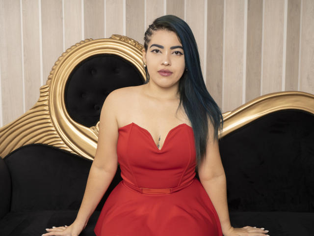 KimLafayette, uma latina curvilínea com mechas azuis, veste um vestido vermelho sem alças, sentada em um sofá preto e dourado, exibindo seus seios e um pequeno piercing no umbigo..