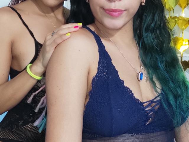 VenusandXiomy with green-blue hair and pink lipstick, wearing a navy lace bra, has her arm touched by another VenusandXiomy in black lace lingerie. Her tits are visible.