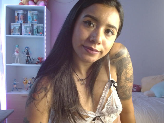 AshleyLover con lunghi capelli scuri, piccoli seni in un reggiseno di pizzo bianco, braccio tatuato, anello al naso e un sorriso discreto, che guarda verso la fotocamera in una stanza illuminata di rosa..