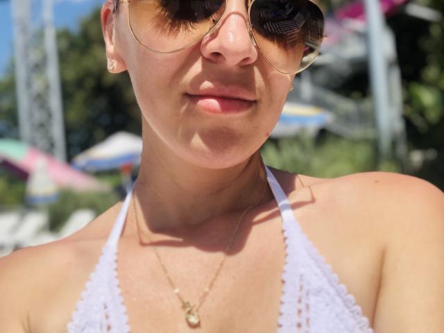 SophieDeee with tan skin, small gold earrings, and a white lace halter top, wearing gold aviator sunglasses, and a gold necklace with a small pendant. She's pouting slightly.