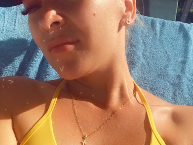 SophieDeee's face with sunlit skin, small droplets on her chin, wearing a yellow bikini top, gold necklace, and small earrings, lying on a blue towel.