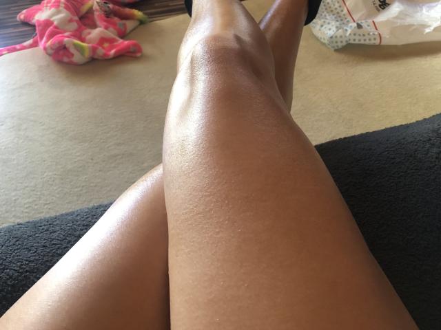SophieDeee's shiny, smooth, brown legs are crossed in the foreground, showing her glistening skin. A pink, fluffy blanket and a white bag are in the background.