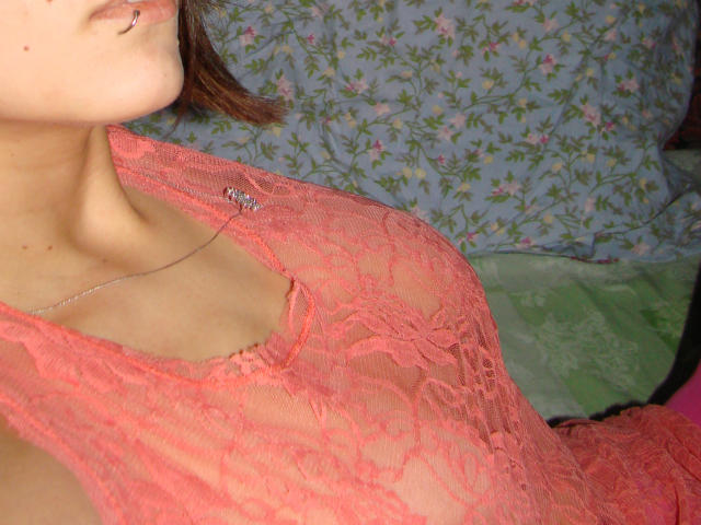 El rostro de RivieraM, con un piercing en el labio, lleva una blusa rosa de encaje transparente que deja ver sus senos, y una delicada gargantilla. Su cabello castaño es corto y apenas visible..