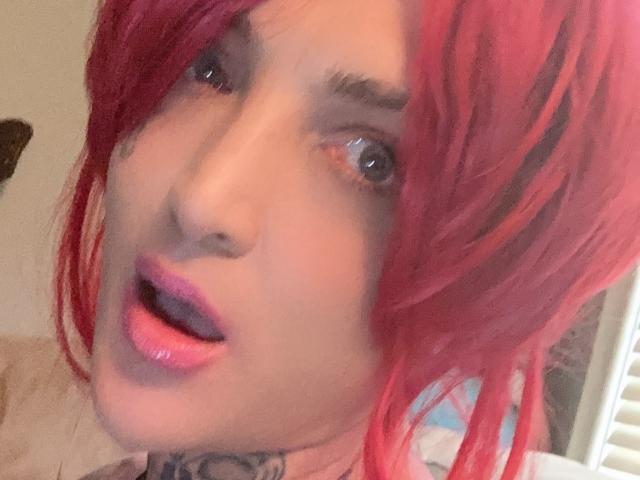 Madelinej hat hellrotes Haar, einen weit geöffneten Mund mit rosafarbenen Lippen und einen überraschten Gesichtsausdruck. Ihre blauen Augen und ihr tätowierter Hals sind sichtbar. Sie blickt nach oben, wobei ihre Zunge leicht herausragt..