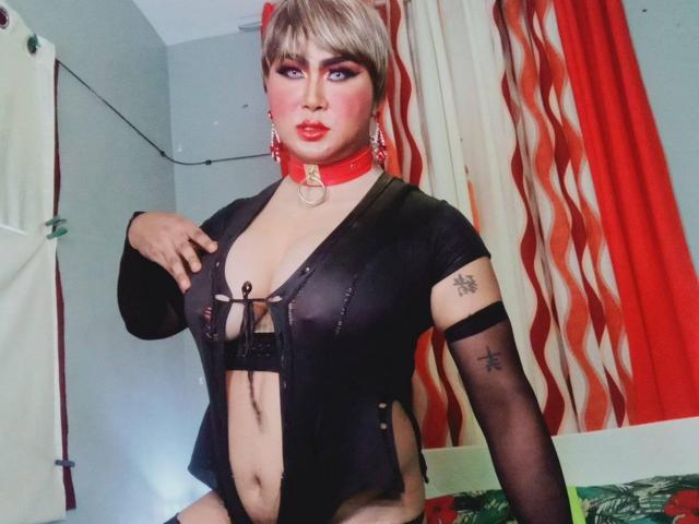 BornStarTS with short blonde hair, red choker, black sheer top, exposed boobs, tattoos on arm, sitting, looking at camera, red and white curtains in background.