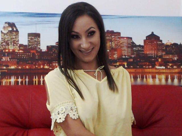 FontainVite, with long dark hair and a smile, wearing a yellow lace-Trimmed top, sits on a red couch, looking cute with her tits slightly visible under the top.