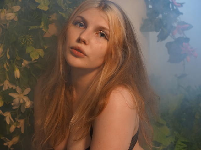 Aleksa Keller ha lunghi capelli biondi, pelle chiara e occhi azzurri, guarda verso l'alto con un'espressione dolce e leggermente curiosa, indossando un reggiseno nero, in un ambiente nebbioso a tema floreale..