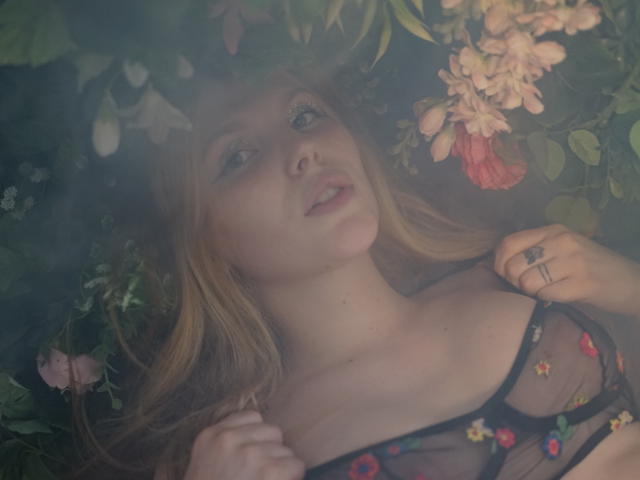 AleksaKeller with blonde hair, wearing a sheer black bra with colorful floral embroidery, lying down, surrounded by flowers, looking up with slightly parted lips, right hand on chest.