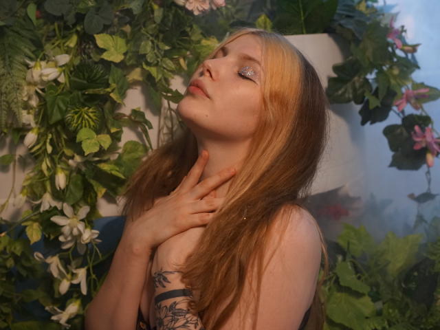Aleksa Keller, con i capelli biondi, un tatuaggio sul braccio destro, gli occhi chiusi, una mano sul collo, un'espressione delicata, circondata da foglie verdi e fiori bianchi, indossa un top nero senza spalline, leggermente inclinato..