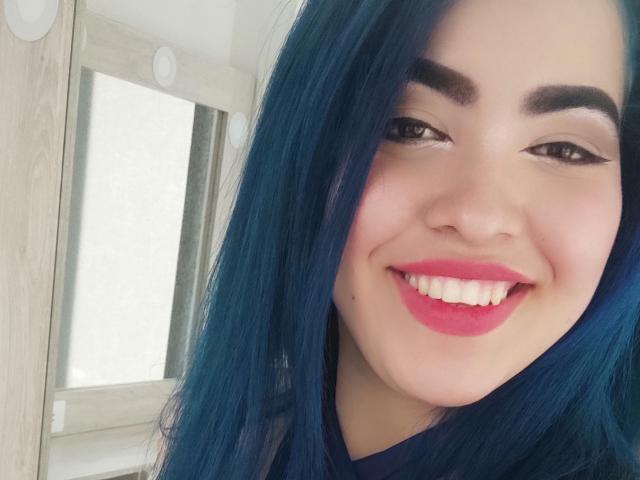 KimLafayette heeft blauw haar, dikke wenkbrauwen en rode lippenstift. Ze glimlacht met witte tanden, draagt een donkere blouse en heeft een schattig, aantrekkelijk gezicht..