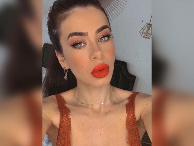 LauraineX has red lipstick, green eyes, and dark brown hair in a ponytail. She's wearing a red lace dress with small earrings and a delicate necklace.
