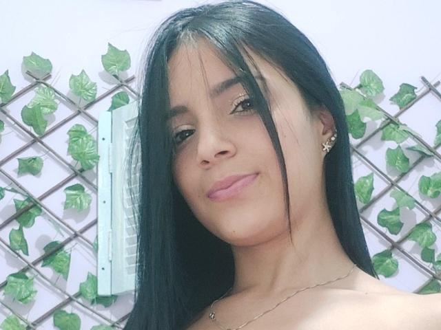 SweetKate69 with long black hair, brown eyes, and pink lips, smiling at the camera. She's wearing small earrings and a thin necklace, with her tits partially visible.