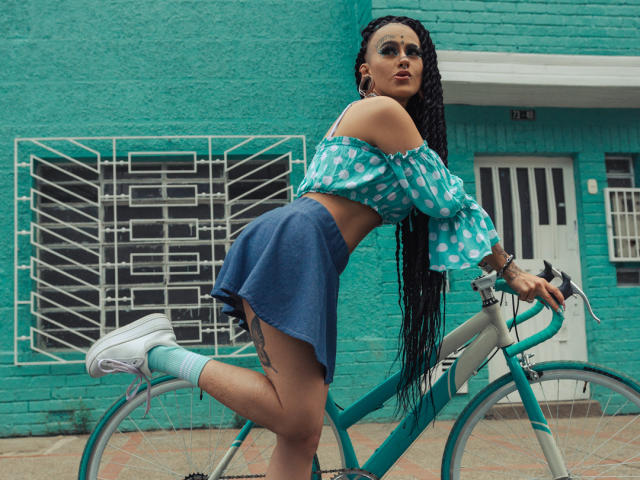 MollyWaxx mit langen Zöpfen, einem blauen, schulterfreien Top, einem Jeansrock, weißen Socken und weißen Turnschuhen, beugt sich über einem türkisfarbenen Fahrrad und zeigt ihr Gesäß, vor einer türkisfarbenen Backsteinmauer..