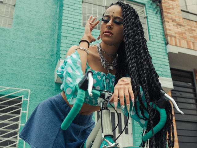 MollyWaxx with long braided hair, wearing a green polka-dot crop top and blue skirt, leaning on a teal bike, multiple necklaces, and dark eye makeup, posing confidently.