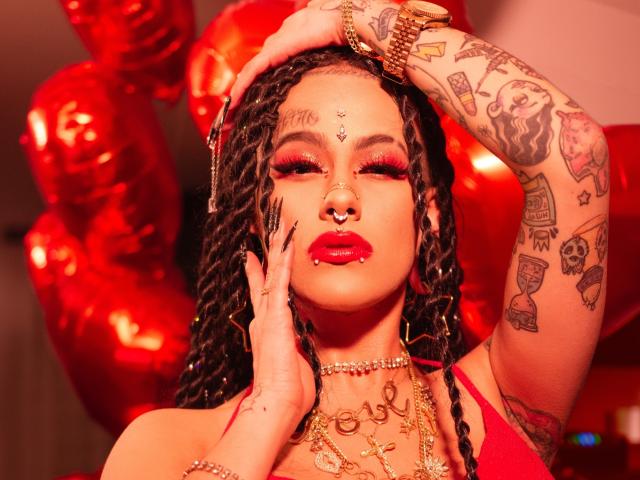 MollyWaxx com batom vermelho, joias douradas e tatuagens, com o cabelo em tranças, balões vermelhos, relógio dourado, piercing no nariz e dedos com anéis, com uma aparência sedutora e olhos fechados..