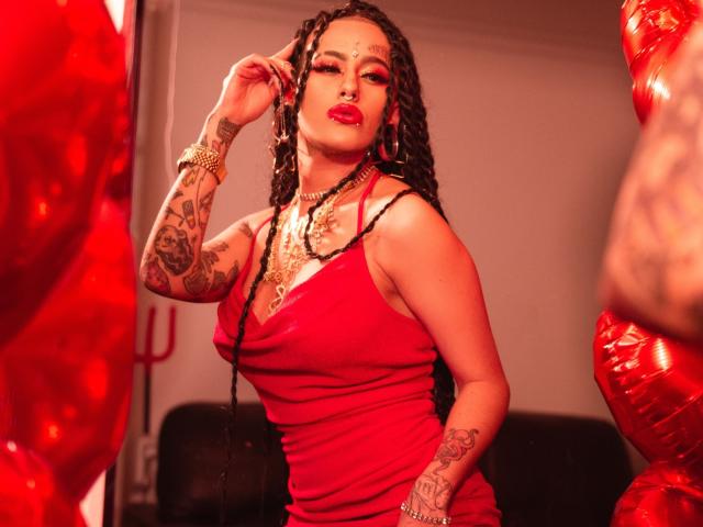 MollyWaxx with long braids, red dress, tattoos, gold necklace, and red lipstick, touching her ear, standing between red balloons, showing her curvy ass and large tits.