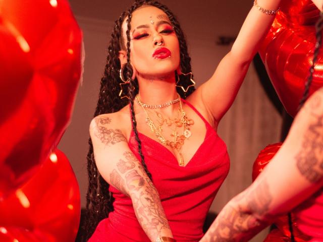 MollyWaxx with long braids, red dress, star earrings, gold necklace, tattoos on arms, red lipstick, nose ring, and red balloons in the background. She's posing confidently.