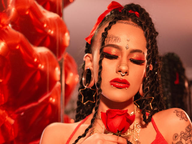 MollyWaxx with red eyes, rose in hand, holding rose to her lips. She's got braids, nose ring, hoop earrings, tattoos, and a red bow. Her tits are covered by a red top.