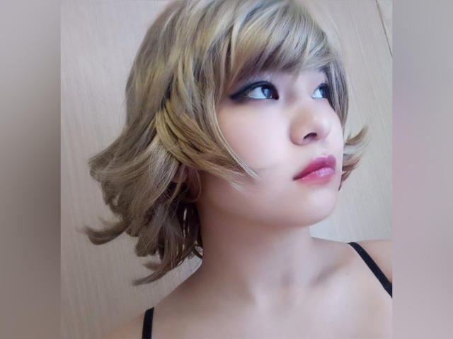 LuxMelys has short, blonde, wavy hair with dark eyeliner, pink lipstick, and black straps on her shoulder. Her skin is pale, and she's looking to the side.