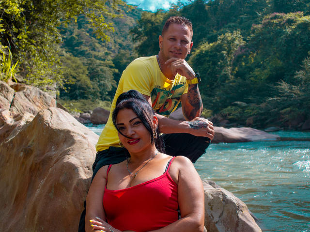 SashaYManuel, mit schwarzen Haaren und rotem Oberteil, zeigt ihre Brüste und sitzt auf Felsen. SashaYManuel mit Tätowierungen, in einem gelben Hemd, kauert im Hintergrund und lächelt. Fluss, grüner Wald, sonnig..