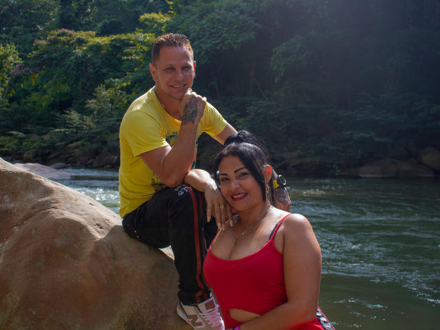 SashaYManuel, com cabelo castanho curto e vestindo uma camisa amarela, está sentada em uma rocha, com as mãos nos joelhos, ao lado de uma mulher curvilínea com cabelo preto e vestindo uma regata vermelha..
