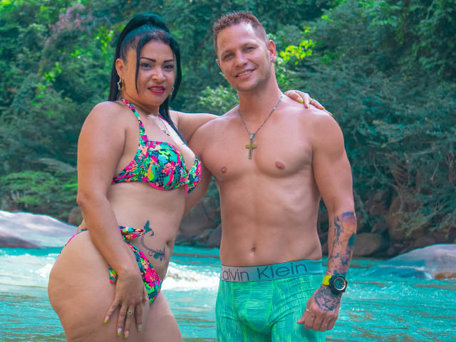SashaYManuel, usando um biquíni colorido, com uma bunda grande e tatuagens, está ao lado de um SashaYManuel sem camisa, vestindo shorts verdes da Calvin Klein, com um colar de cruz e tatuagens, em um cenário tropical com água..