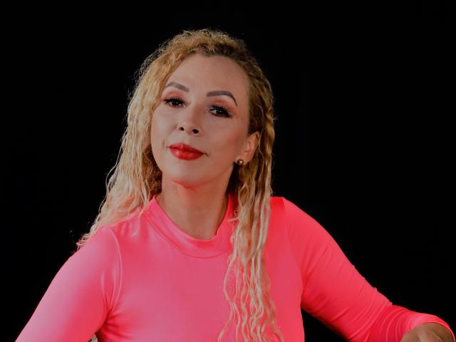 SandyFit with wet blonde curly hair, pink long-sleeve top, red lipstick, and earrings, smiling against a black background.