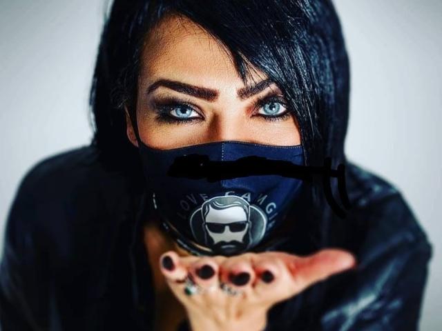 CharlotteJhons, met zwart haar, blauwe ogen en een zwart masker dat haar neus en mond bedekt, steekt een hand uit met zwarte nagels, en draagt een zwarte leren jas..