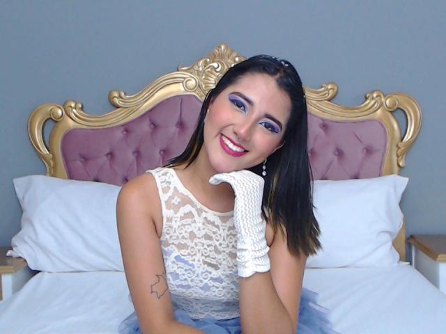 AgathaRose, aux yeux bleus, souriante, portant une robe blanche en dentelle, des gants blancs et du fard à paupières violet, assise sur un lit avec une tête de lit rose et dorée, montrant légèrement ses seins..