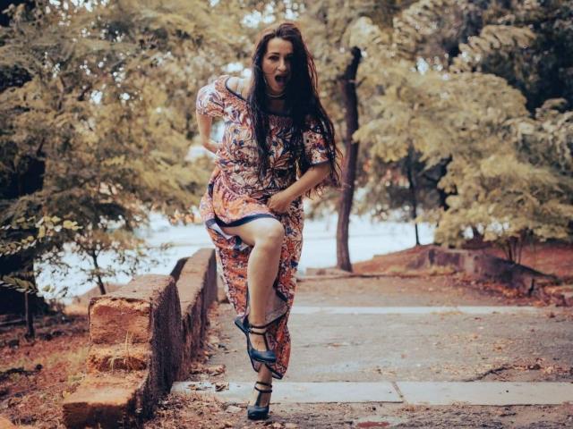 GinaONeon with long brown hair, colorful patterned dress, and black heels, running with one leg raised, mouth open in surprise, on a stone pathway with trees.