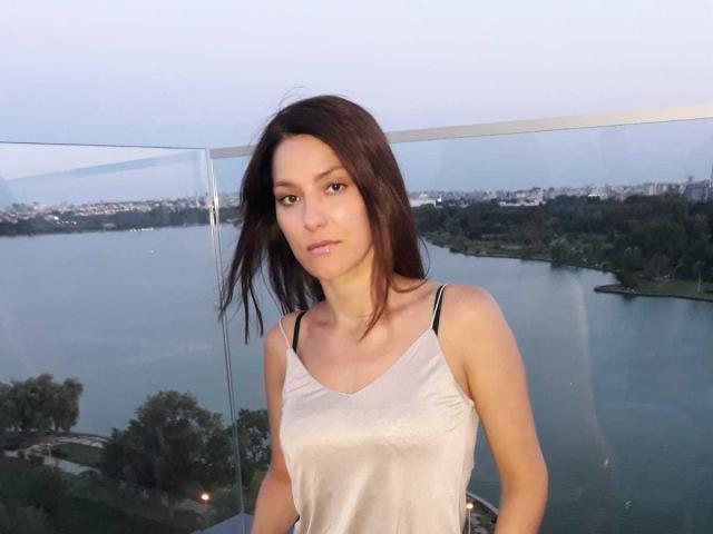JoliDanielle has medium-length brown hair, a serious face, and is wearing a white spaghetti-strap top, showing her small tits and slim arms. She stands by a glass railing.