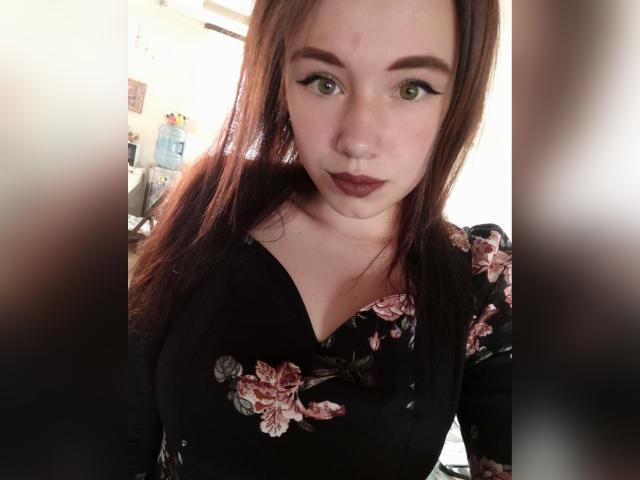 KiraNightly, met lang bruin haar, groene ogen en donkere lippenstift, draagt een zwarte jurk met bloemenprint, waarbij haar borsten zichtbaar zijn, en kijkt met een licht verraste blik in de camera..