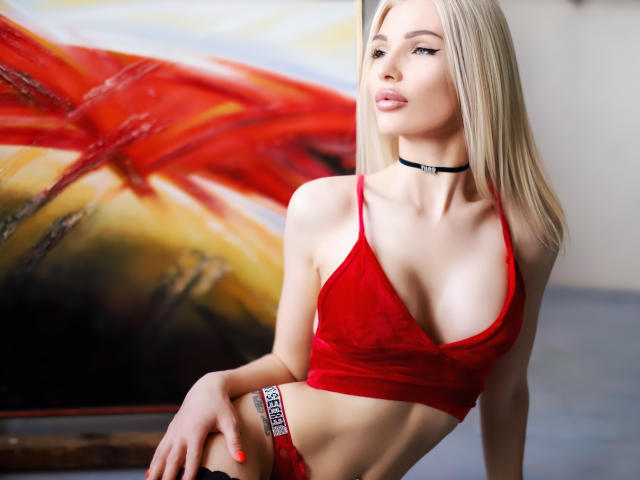 PleasingAngel with blonde hair, red velvet bra, choker, and red lace panties, showing her small tits and flat ass, standing in front of a colorful abstract painting.