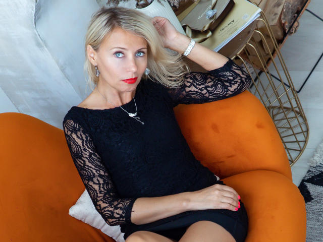 OneLadySexy with blonde hair, blue eyes, red lipstick, wearing black lace top, sitting on orange chair, hand on head, black skirt, white bracelet, looking at camera.