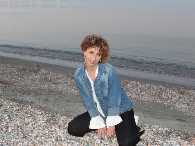 NikHotty, con i capelli castano ricci, una giacca di jeans blu, una camicia bianca e pantaloni neri, è inginocchiata su una spiaggia di ciottoli, sorride alla fotocamera, mostrando il suo piccolo sedere e il suo viso carino..