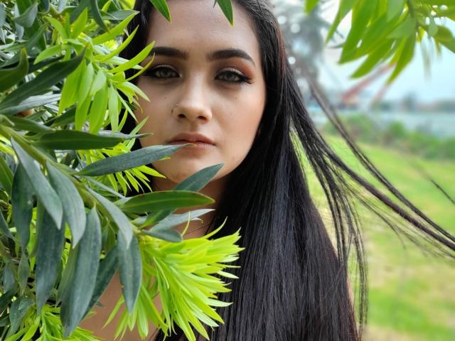 EmmaTayllor with long black hair, green eyes, and subtle makeup, hiding behind green leaves with a focused expression, her nose slightly visible, in a lush outdoor setting.