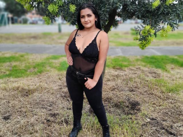 EmmaTayllor è in piedi all'aperto, indossa lingerie in pizzo nero, una maglia trasparente e jeans neri aderenti, con un albero con foglie verdi sopra di lei, sorride, mostrando seni di medie dimensioni e un piccolo tatuaggio sul petto..