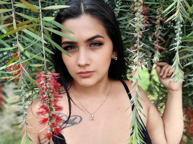 EmmaTayllor, con cabello negro, ojos verdes y un maquillaje sutil, está de pie entre ramas verdes y rojas, vistiendo una blusa negra con un pequeño tatuaje en el pecho..