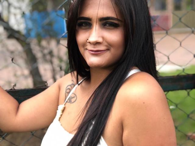 EmmaTayllor, con cabello negro largo, sonriendo, con una camiseta blanca sin mangas, un piercing en la nariz y un tatuaje pequeño en el hombro derecho, de pie junto a una cerca de alambre, sosteniéndola con su mano derecha..