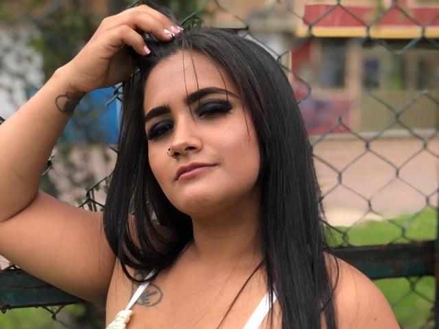 EmmaTayllor, con cabello negro, maquillaje de ojos oscuro, un piercing en la nariz y un tatuaje, levantando la mano, vistiendo una blusa blanca, luciendo segura de sí misma, de pie frente a una valla de alambre..