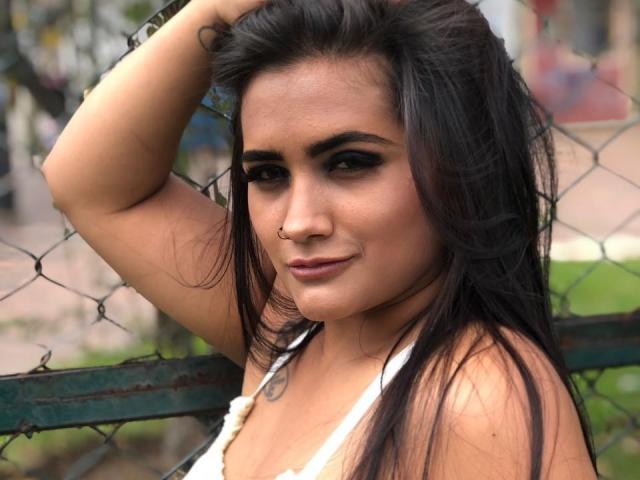 EmmaTayllor with long black hair, nose ring, dark eye makeup, white halter top, hand on fence, slight smile, tan skin, green background, subtle tattoo on left arm.