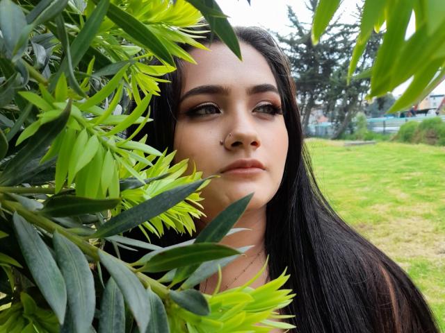 EmmaTayllor with long black hair, green eyes, and subtle makeup, partially hidden by bright green leaves, looking off-camera with a calm expression. Her nose is in the center.