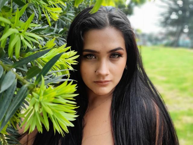EmmaTayllor with long black hair, green eyes, and a serious expression, standing near bright green leaves, showing a subtle nose ring and a calm, sexy look.
