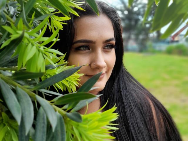 EmmaTayllor, con lunghi capelli neri, occhi verdi, un anello al naso e un sorriso discreto, è parzialmente nascosta da foglie verdi, in un ambiente esterno. Le sue natiche non sono visibili..