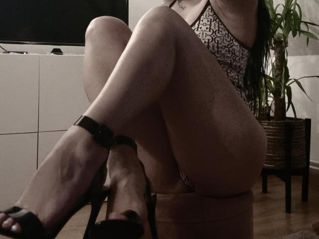 Belisa's legs crossed, wearing a leopard-print lingerie set, black high heels, and her bare ass is visible. Her long black hair falls over her shoulders. Dimly lit room.