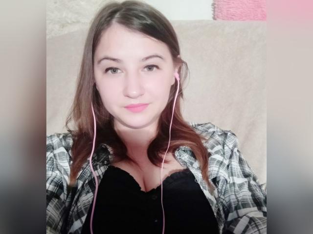 MiraDickey mit rosa Kopfhörern, die einen schwarzen BH, ein kariertes Hemd und pinken Lippenstift trägt, hat eine mittelgroße Brust und braunes Haar und lächelt in die Kamera..