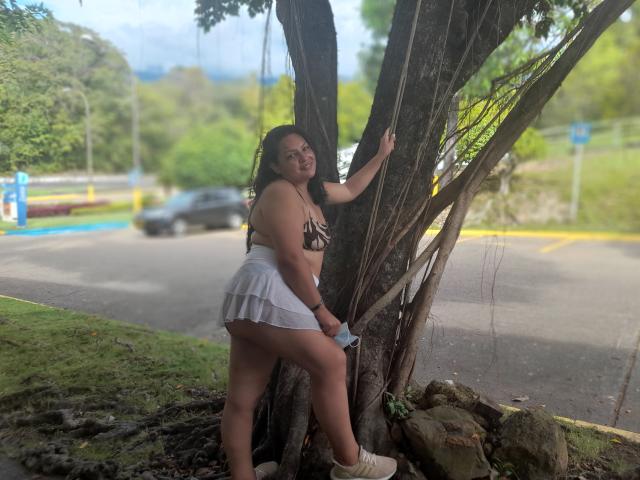 Adi69, avec sa peau brune et ses longs cheveux noirs, se tient contre un arbre, vêtue d'un soutien-gorge léopard et d'une mini-jupe blanche plissée, montrant ses fesses et tenant un téléphone..
