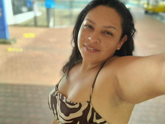 Adi69 met middelgrote borsten, een bruin-witte gestreepte bikini top, lachend, zwart haar, arm omhoog, oksel zichtbaar, buiten, daglicht, lichte gebruinde huid, casual selfie..