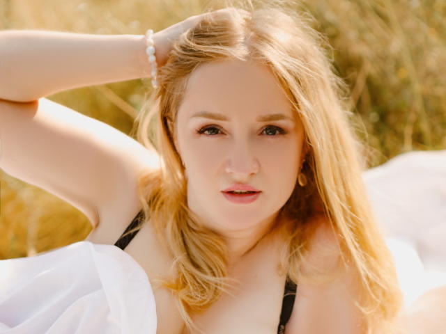 OhMollyHoney with blonde hair, fair skin, and pink lips, wearing a black strap top, hand in hair, white cloth, and a pink bracelet, looking directly at the camera.