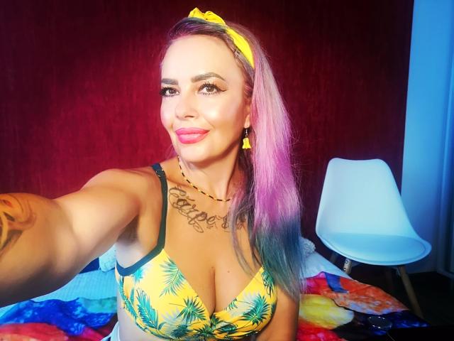 HotAdelayde69 with pink and purple hair, yellow headband, yellow pineapple bra, tattoos, and pink lipstick, taking a selfie, showing her big tits and smiling.